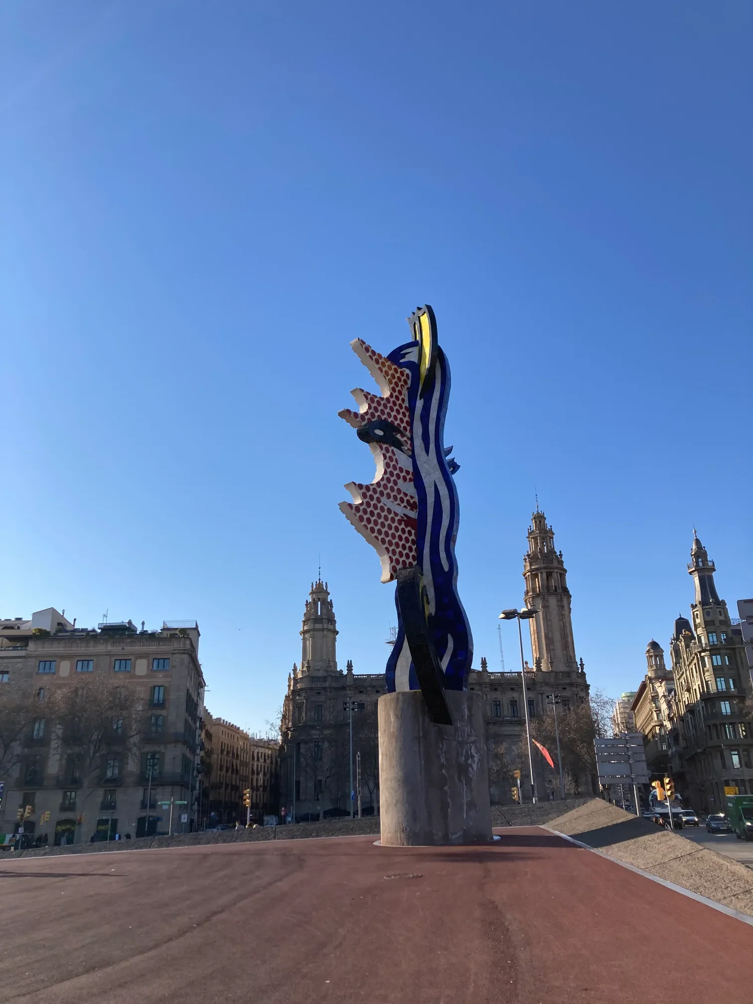 Kolorowa rzeźba El Cap de Barcelona autorstwa Lichtensteina przy nadbrzeżu Barcelony z zabytkowymi wieżami w tle