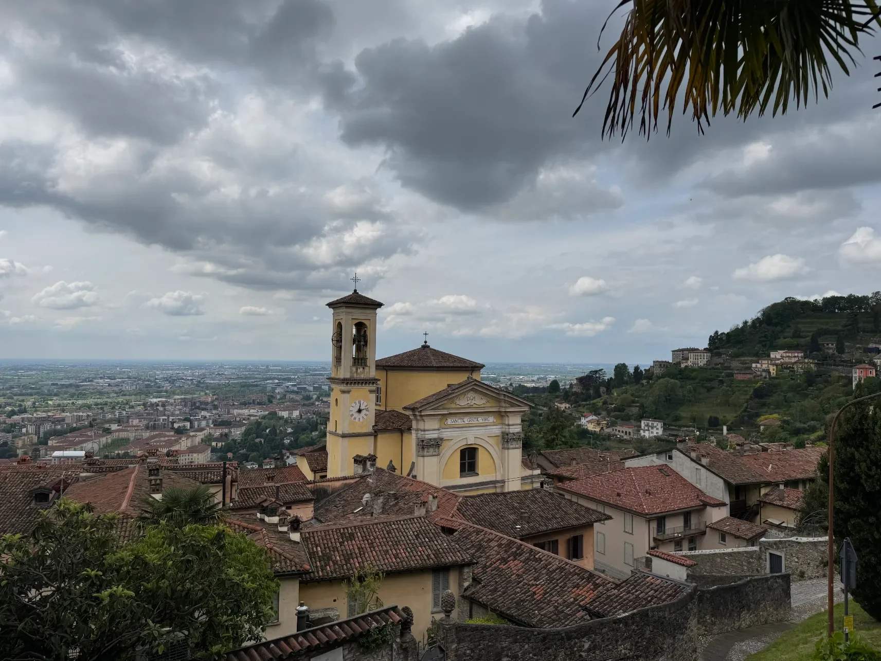 Panorama Bergama se střechami Citta Alta a žlutým kostelem s hodinovou věží na pozadí lombardských rovin