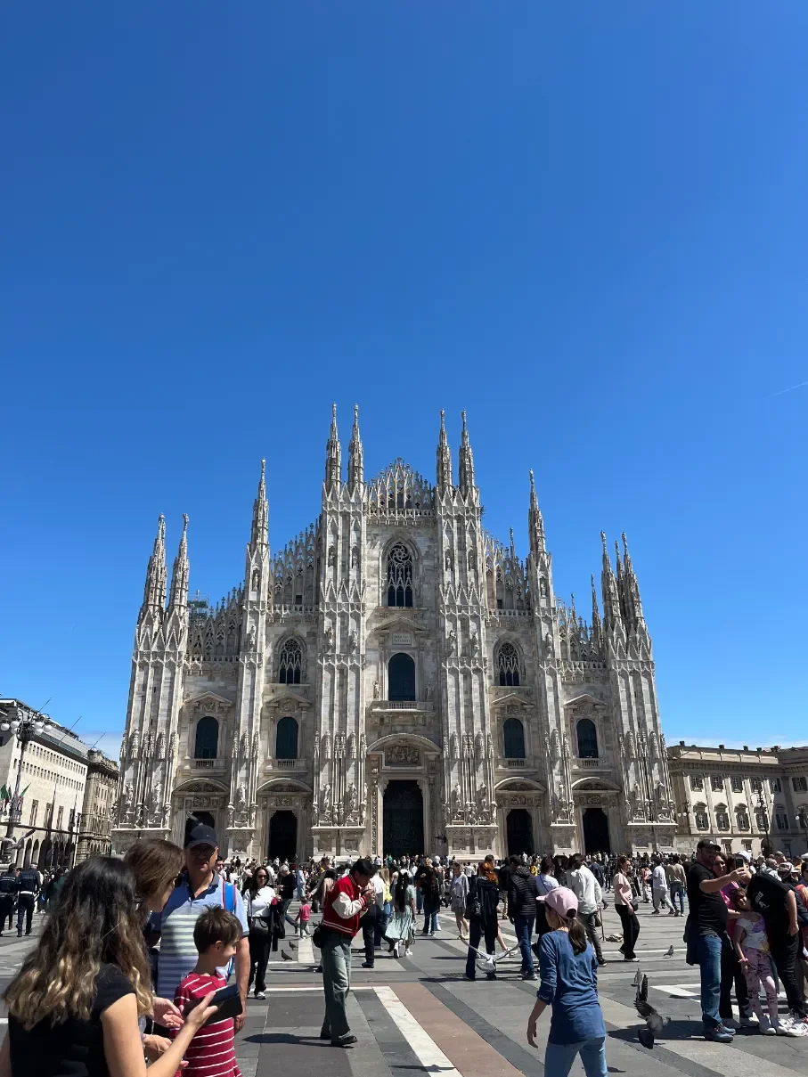Gotická fasáda katedrály Duomo di Milano s mramorovými fiálami a davem turistů na Piazza del Duomo