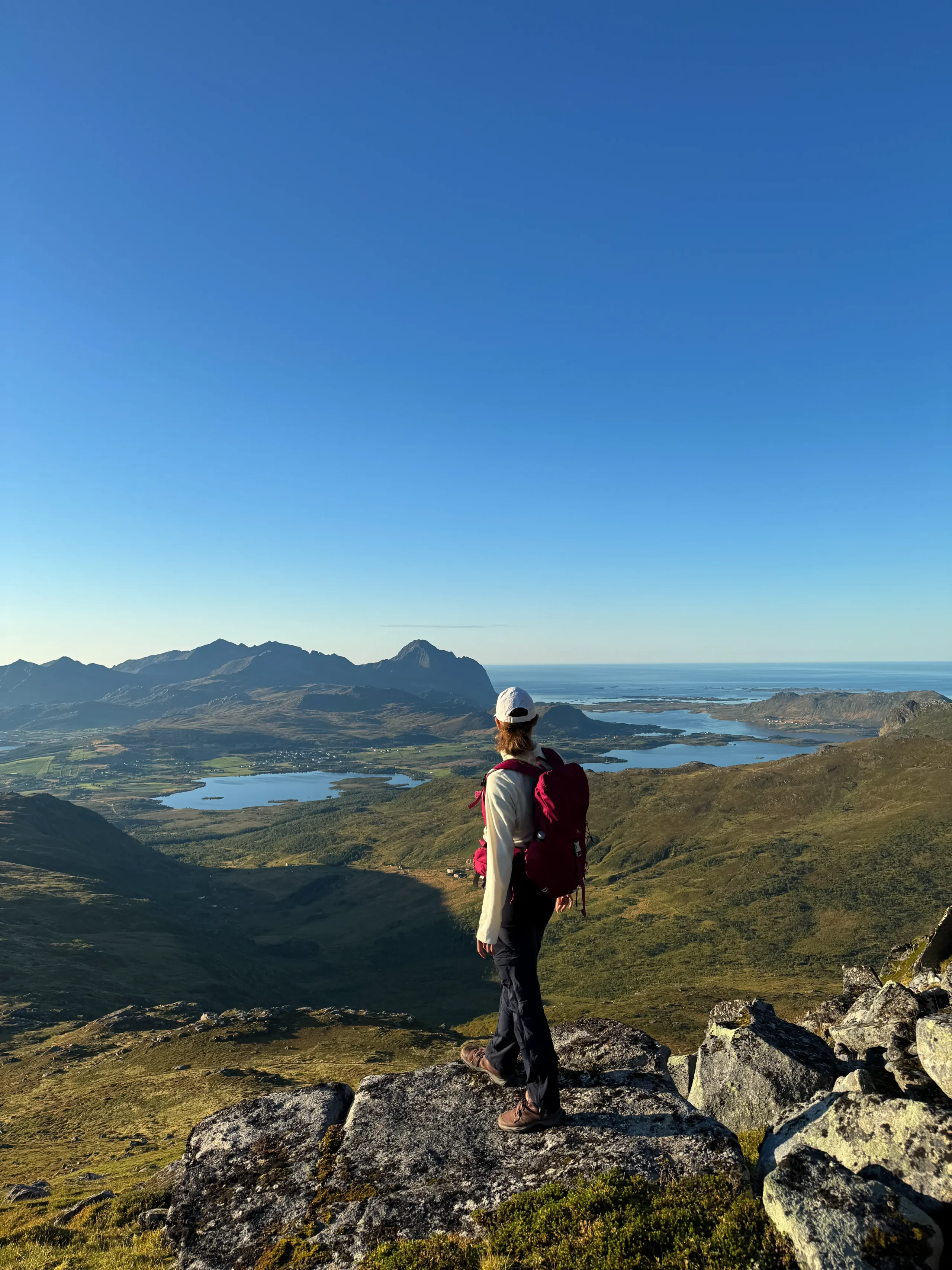 Turistka s červeným batohem stojící na skalním hřebeni Blåtinden a obdivující panorama ostrova Vestvågøya s fjordy na Lofotech