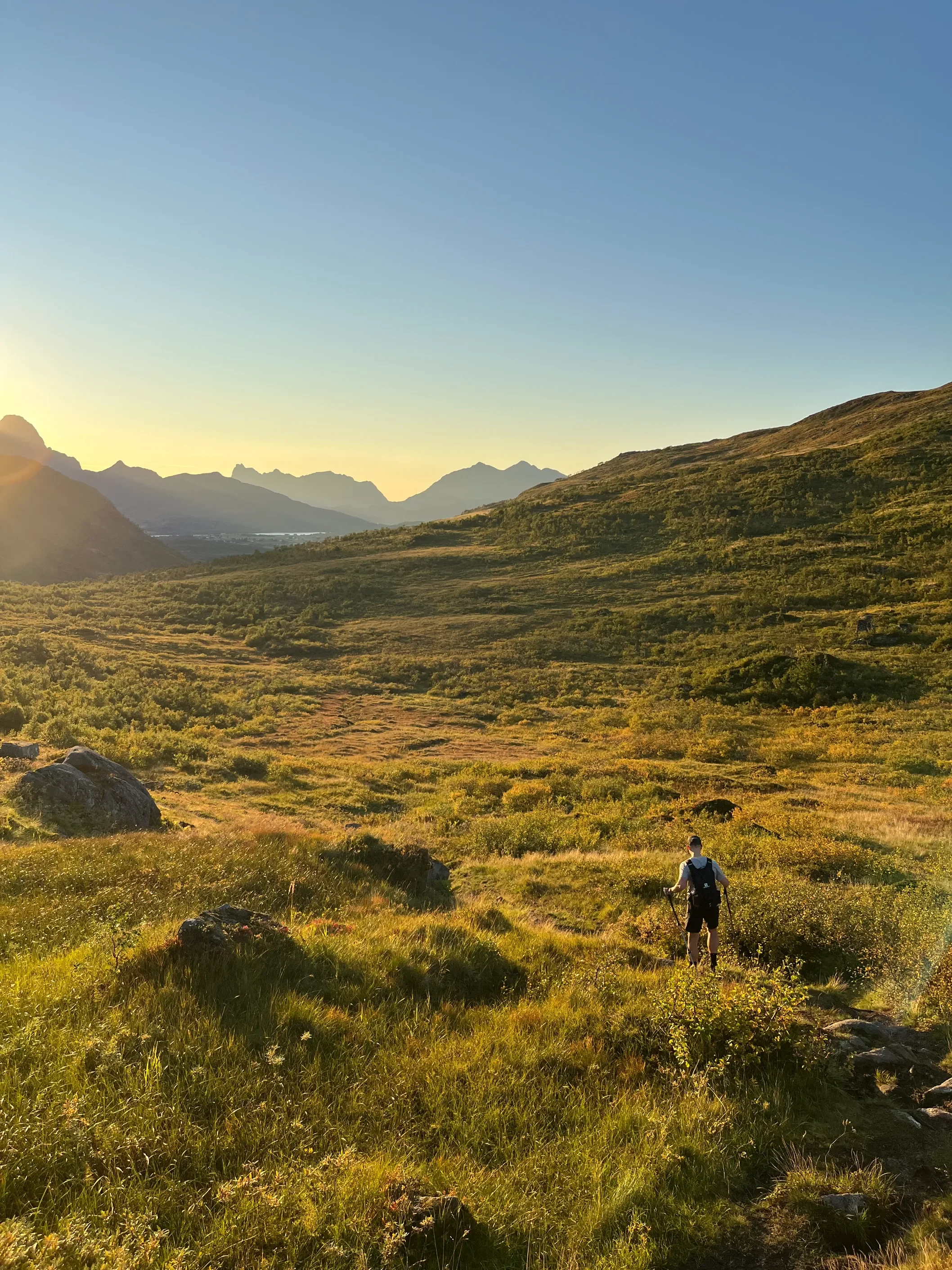 Turista s hůlkami na rozlehlé zelené plošině trasy Blåtinden ve zlaté hodině se siluetami vrcholů Lofot a fjordy v pozadí
