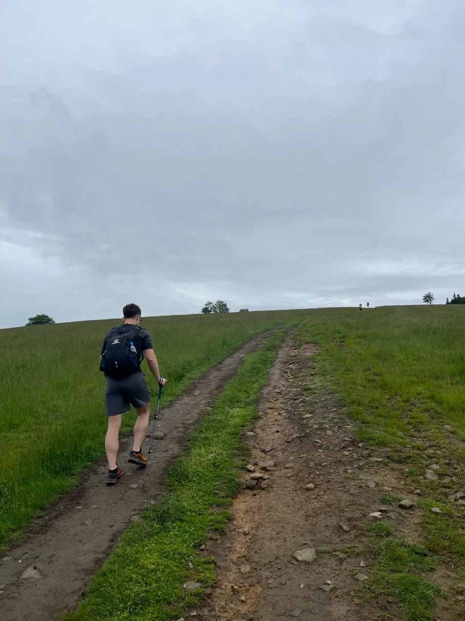 Hiker with trekking pole climbing a wide path across the open meadow toward the Hala Jaworowa clearing on Kotarz