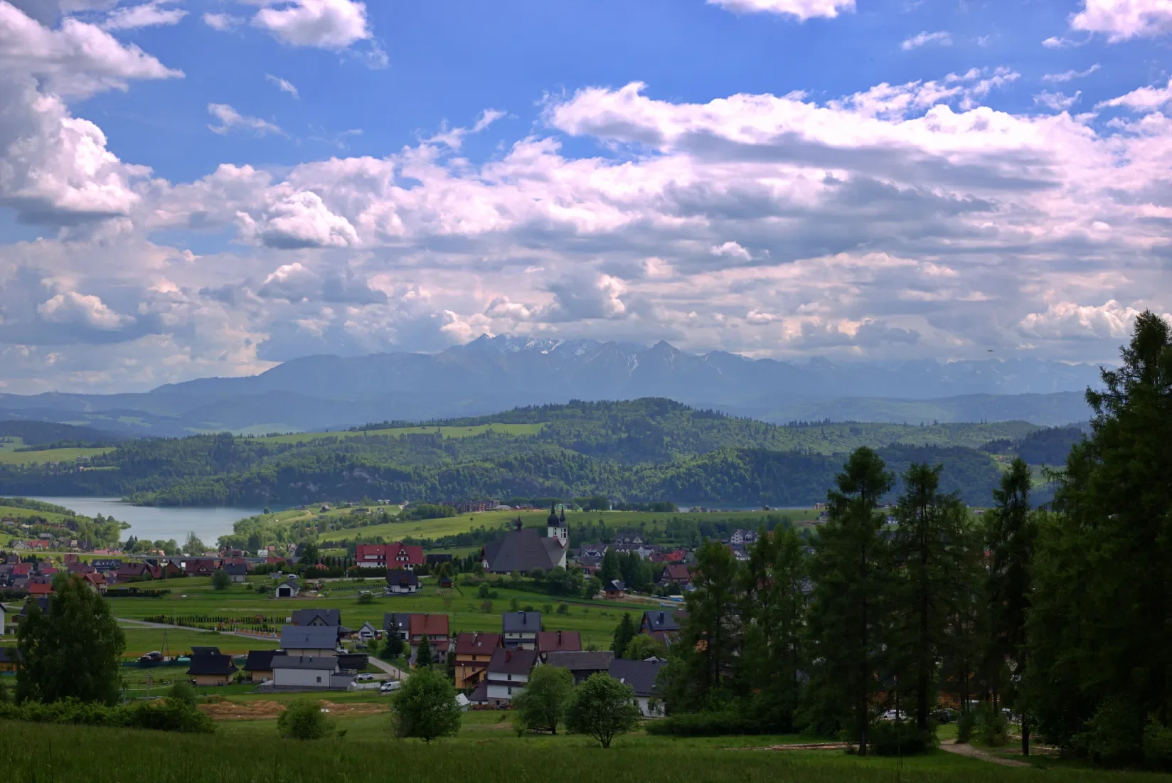 Výhled ze žluté značky na podhalskou vesnici s kostelem, Czorsztyńské jezero a zasněžené Vysoké Tatry na obzoru