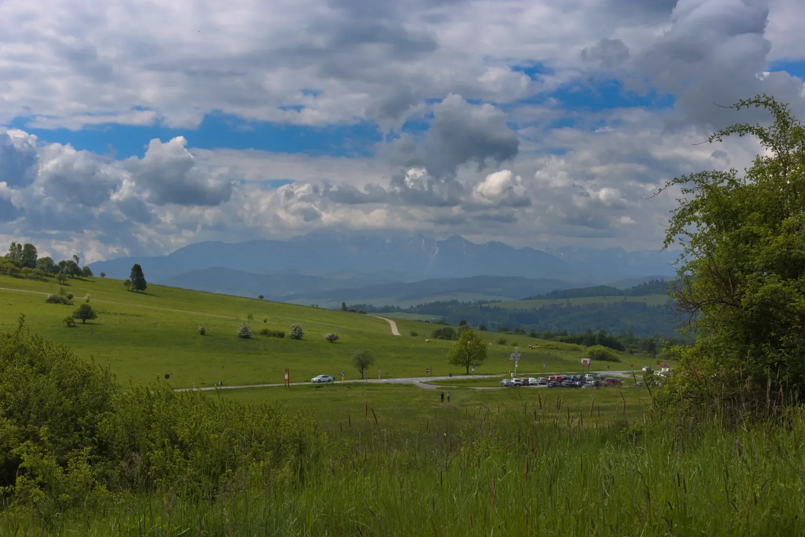 Zelená louka klesající k parkovišti, zamlžené Vysoké Tatry na obzoru a dramatické mraky, jaro