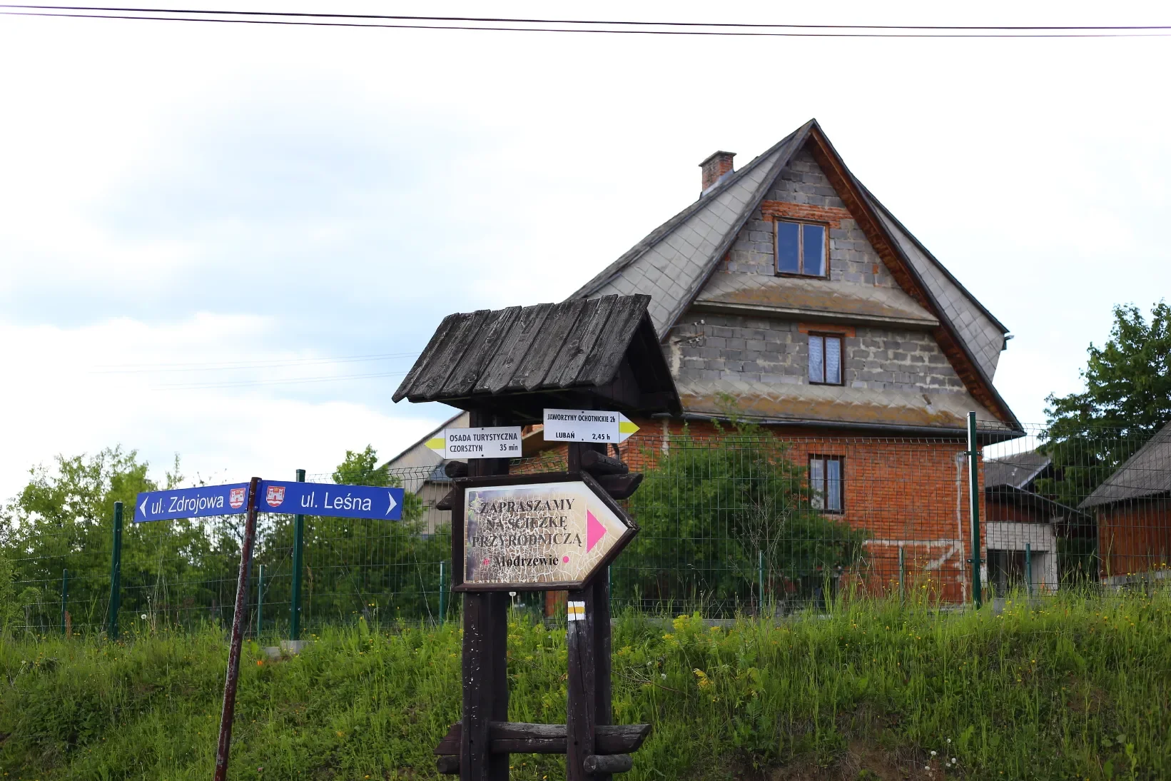 Vesnický rozcestník s tabulkami do Czorsztyna a na Lubań (2:45) vedle zděného domu, začátek naučné stezky