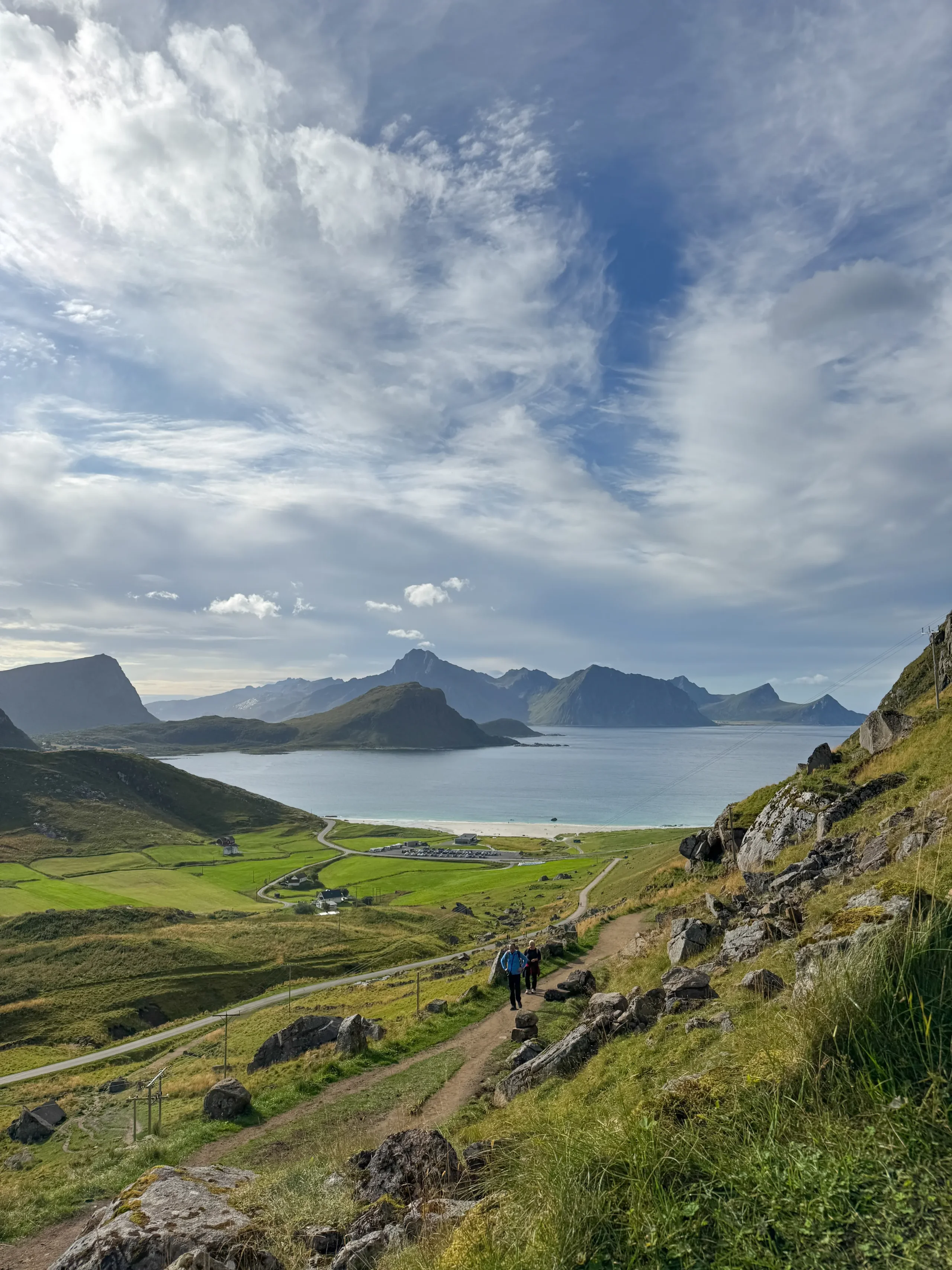 Turisté sestupující strmou trasou z Mannenu s výhledem na pláž Haukland a lofotské vrcholy
