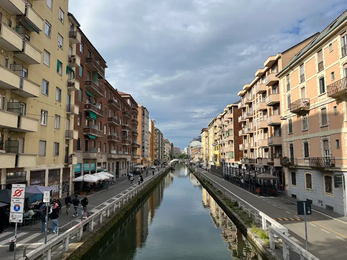 Kanál ve čtvrti Navigli v Milánu, domy po obou stranách vody a kavárny se slunečníky pod zataženou oblohou