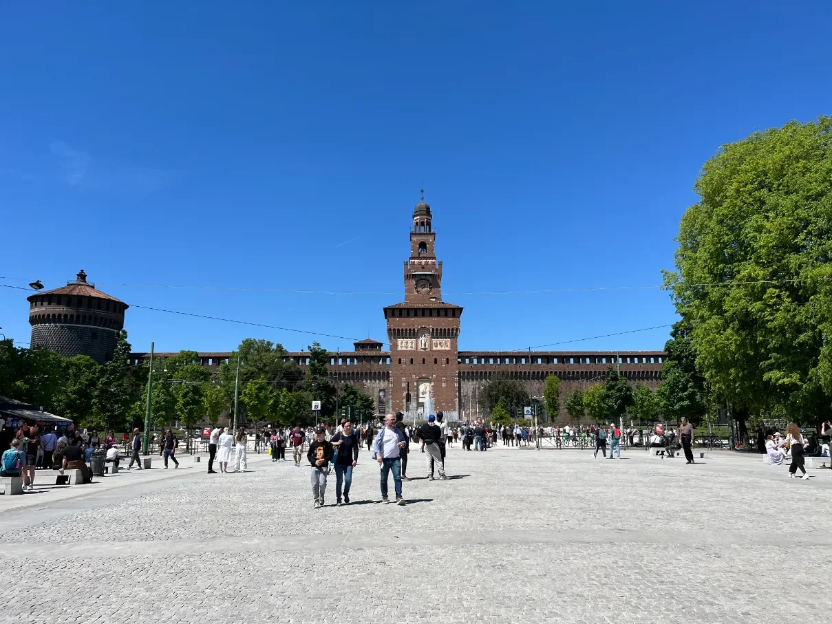 Cihlová pevnost Sforzovského hradu (Castello Sforzesco) s hodinovou věží a nádvořím plným turistů za slunečného dne, Milán