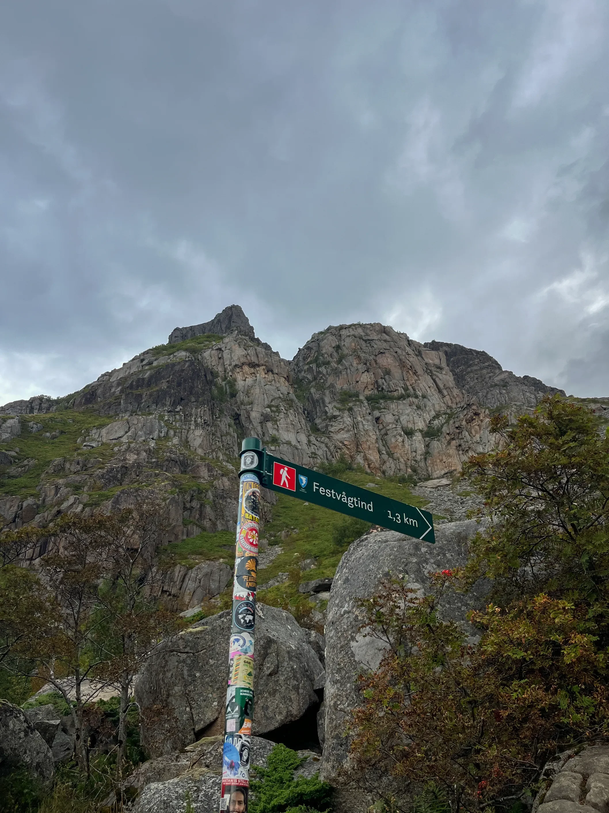 Rozcestník Festvågtind 1,3 km se sloupkem polepeným nálepkami na pozadí skalnatého vrcholu Festvågtind, Lofoty