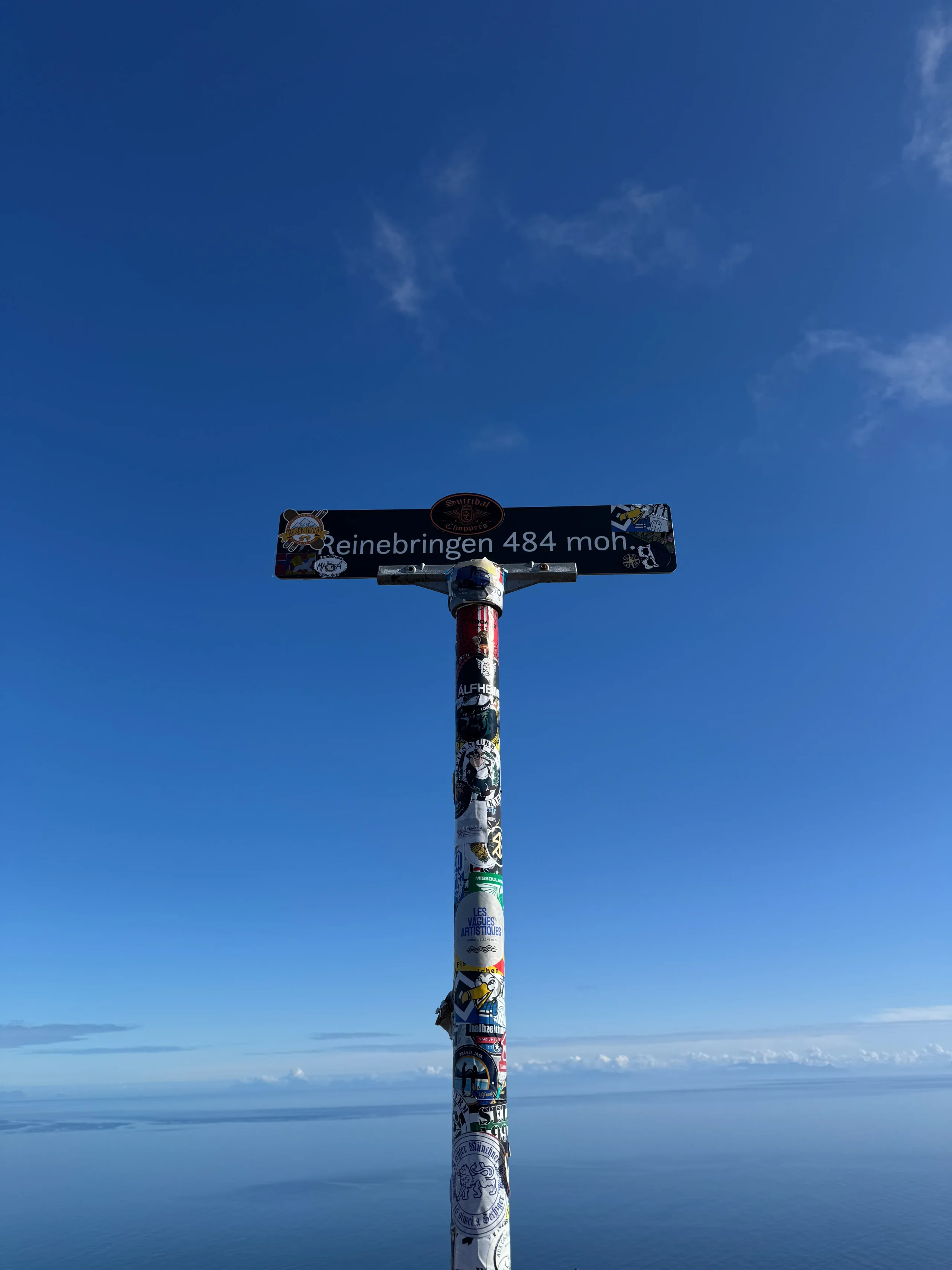 Nálepkami polepený vrcholový sloupek s tabulkou „Reinebringen 484 moh“ na pozadí modrého moře a nebe