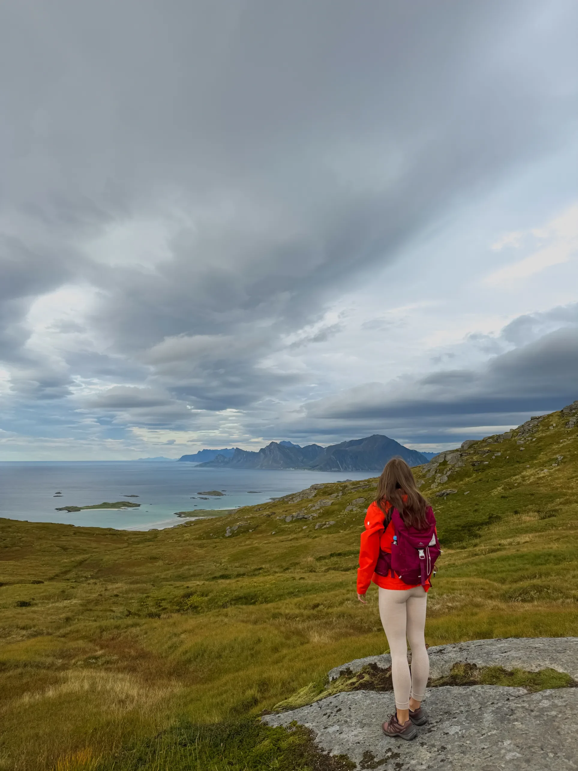 Turistka v červené bundě na kamenitém svahu trasy Ryten s panoramatem fjordu a vrcholů Lofot
