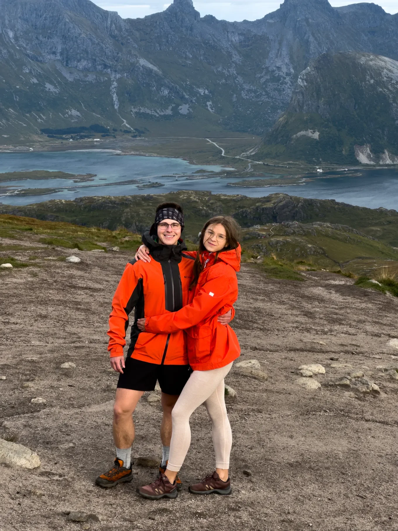 Dvojice turistů v oranžových bundách na vrcholu Ryten s rozlehlým panoramatem fjordu a hor Lofot