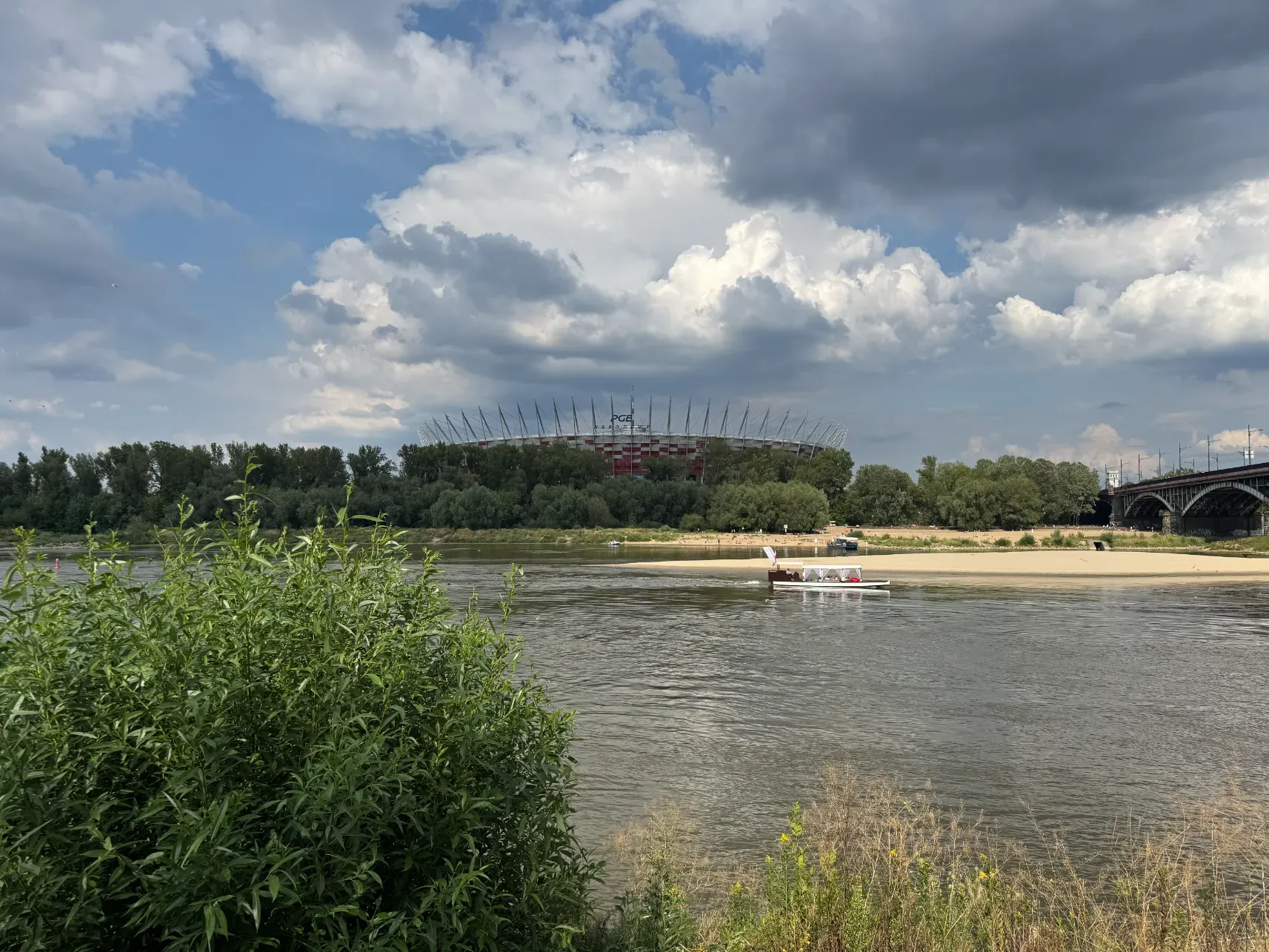 Stadion Narodowy PGE widziany zza Wisły, piaszczysta plaża, mała łódka i letnie chmury nad rzeką